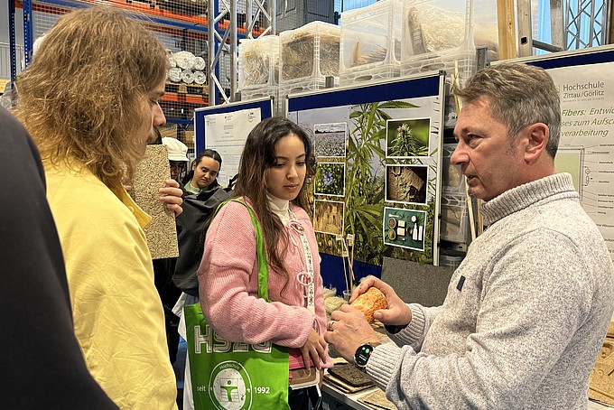 Matthias Tirsch stands in the LANDER³ hall of the HSZG and shows a group of visitors various sustainable materials. Information posters and stored materials can be seen in the background. The participants listen attentively and look at the objects on display while Tirsch explains the innovative application possibilities and the contribution of this research to environmental protection.