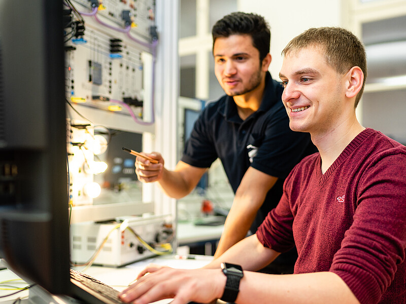 Zwei Studenten im Elektrotechniklabor, einer sitzt am Rechner, der andere steht dahinter und deutet auf den Bildschirm.