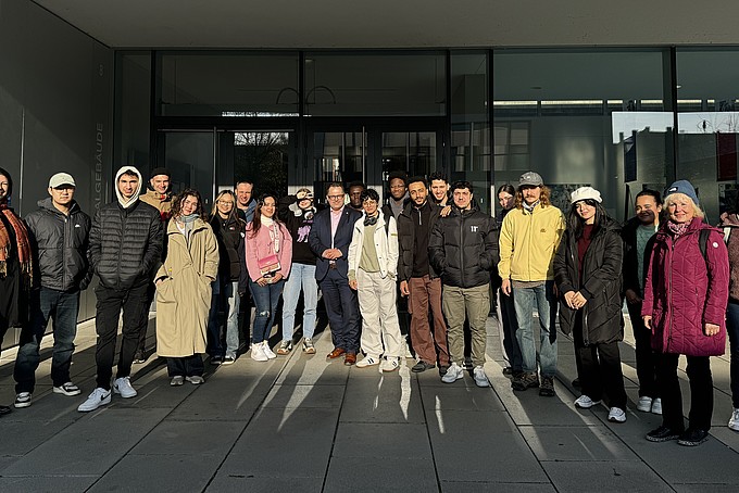 A group of international prospective students from the Rahn Studienkolleg Leipzig pose for a group photo together with the Rector of the HSZG in front of the entrance area of the university. Everyone is standing close together and smiling at the camera as the afternoon sun illuminates the entrance to the building.