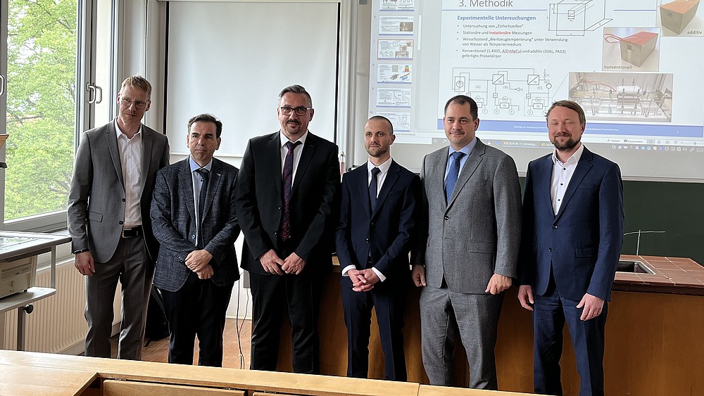 In a seminar room in front of a projector screen with a presentation, the participants in the doctoral defense stand in a row for a group picture.