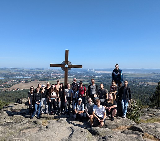 A group photo of a student hiking group in the Zittau Mountains in front of a summit cross under a blue sky.