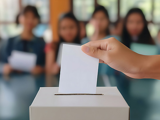 Jugendliche sitzen im Hörsaal im Hintergrund, vor Ihnen sieht man eine Hand, die einen weißen Zettel in eine Wahlurne einwirft. 