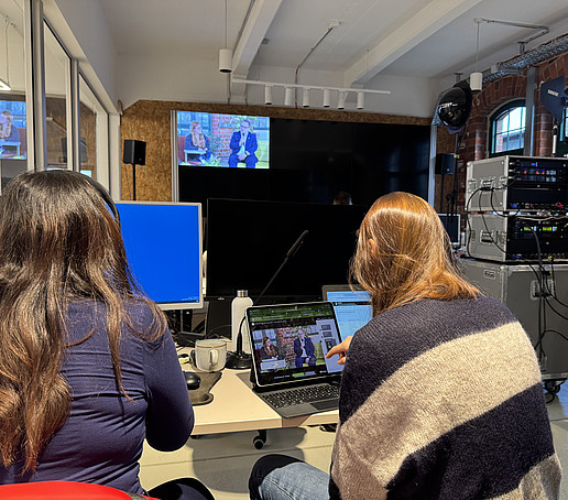 In a room separated from the studio by a glass wall, two employees sit in front of screens and look at them. A large screen can be seen in the background.