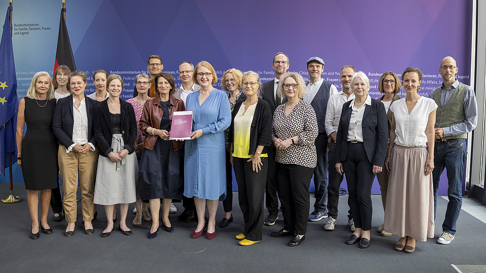 Advisory Board members and the Minister for Family Affairs in front of a press wall in the Bundestag stand next to each other for a group photo. The Minister holds the report in her hand.