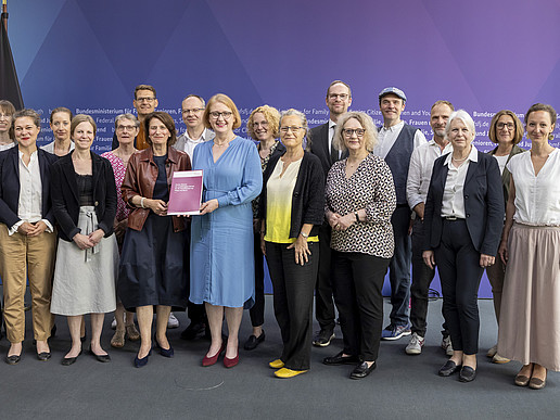 Advisory Board members and the Minister for Family Affairs in front of a press wall in the Bundestag stand next to each other for a group photo. The Minister holds the report in her hand.