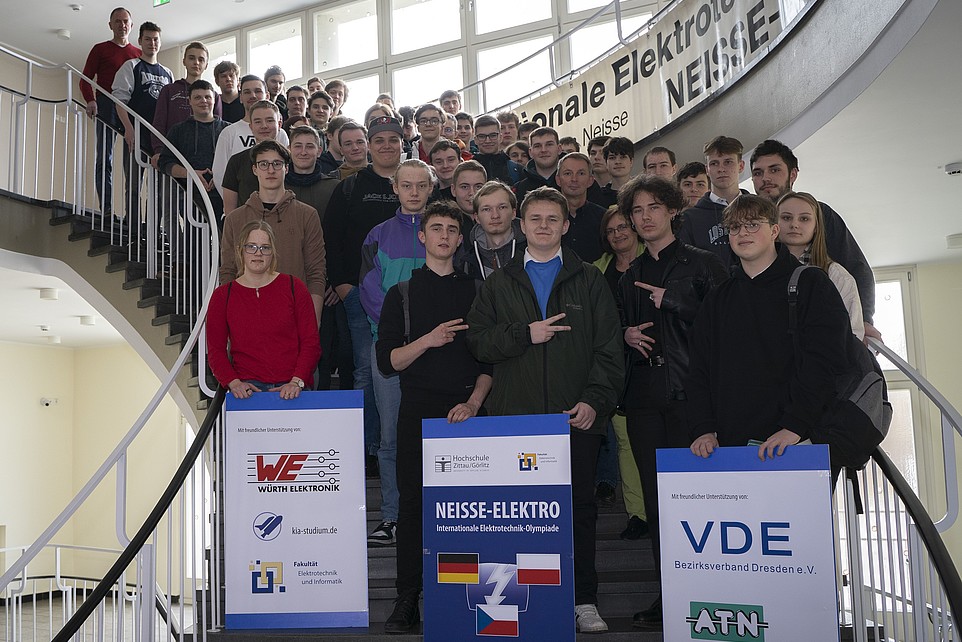 A group of students stands on a staircase. The ones in front are holding three posters with the advertising partners and the logo of Neisse Elektro
