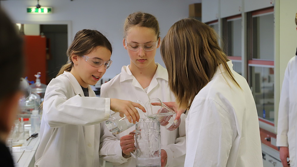Drei Schülerinnen in weißen Kitteln experimentieren im Chemielabor.