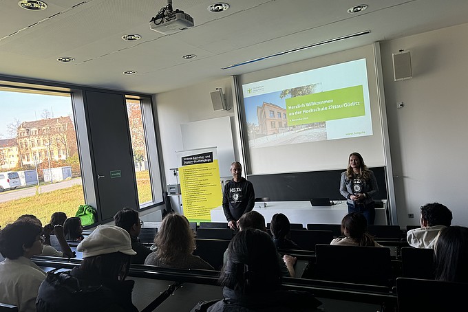 Robert Viertel and Inna Hauf stand at the front of a lecture hall and speak to prospective international students at the Rahn Studienkolleg Leipzig. A presentation entitled "Welcome to Zittau/Görlitz University of Applied Sciences" can be seen in the background. The audience sits in rows and listens attentively to the welcome.