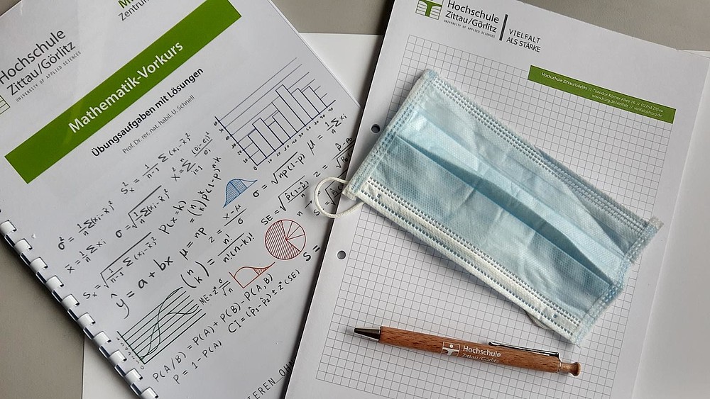 On the left side you can see the title page of the exercises for the math pre-course, on the right side a checkered writing pad with the university logo. On the pad is a typical medical disposable mouth and nose protector and a wooden ballpoint pen with the university logo.