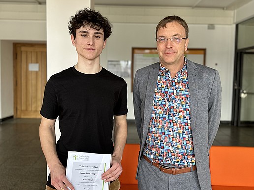 Tom Saugel with certificate in his hands and Prof. Kroschel standing in the hallway of a university building.