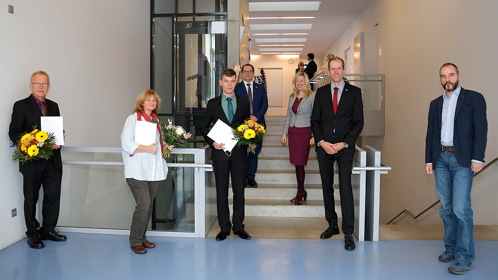 A group of people are standing in the stairwell. Three are holding bouquets of flowers and certificates. They are all smiling at the camera.