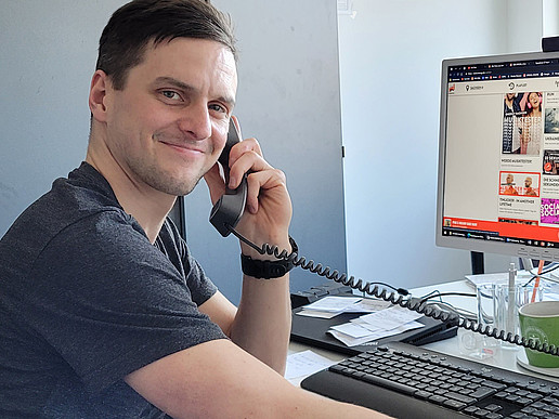 Rico Winkler smiles into the camera, sitting at his desk with a telephone receiver in his hand and the Radio Energy Sachsen website displayed on his PC screen in the background.
