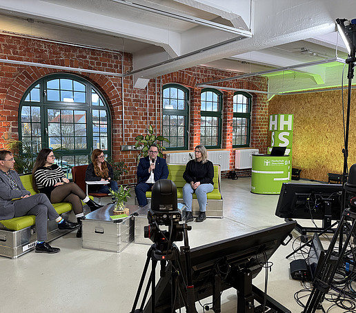 In a studio with brick walls and large windows in the background, participants sit on green sofas. There are black screens on the floor in the foreground. There is a light screen at the edge.