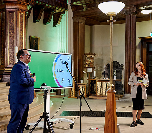 Rektor vor dem Buzzer, im Hintergrund ein Bildschirm mit digitalem Glücksrad, rechts steht die Moderatorin.