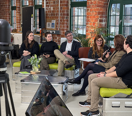 In a studio with brick walls and large windows in the background, participants sit on green sofas and chat.