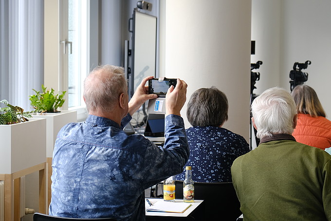 Im Seminarraum filmt ein Senior mit seinem Smartphone einen Vortrag.