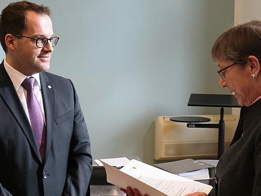 A middle-aged man with a purple tie and black suit, wearing a white shirt underneath and glasses, is presented with a certificate by a woman. A pastel blue-green wall can be seen in the background.