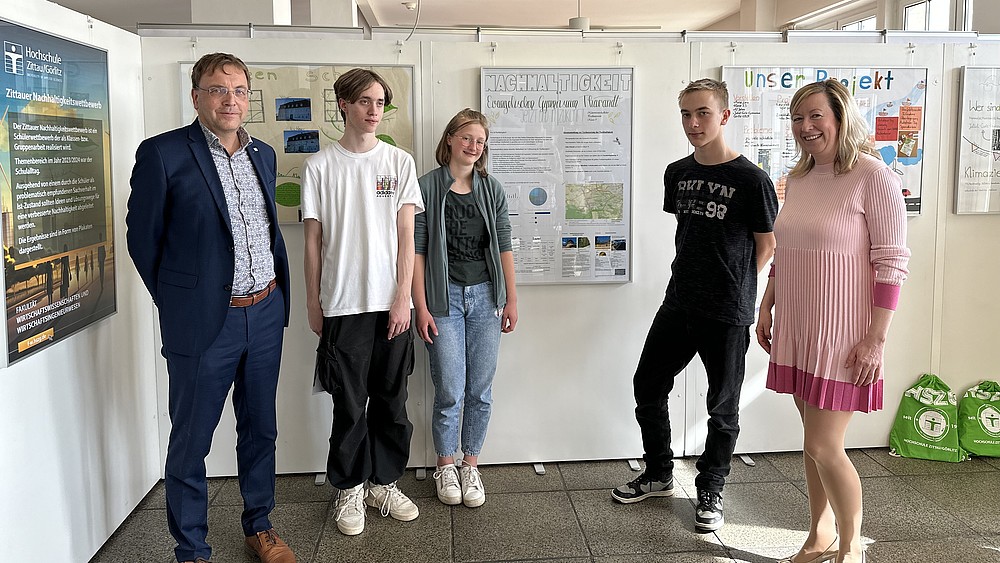 The dean, two students, one student and the vice-rector stand in front of white back walls with posters on the topic of sustainability pinned to them and smile into the camera.