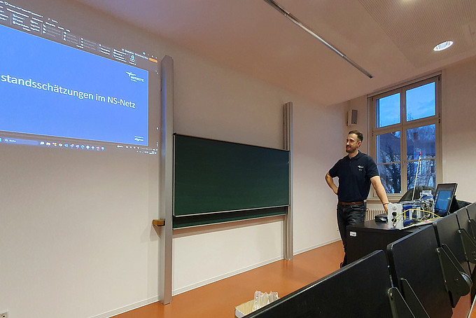 A man dressed in black with a beard is giving a presentation in a lecture hall.