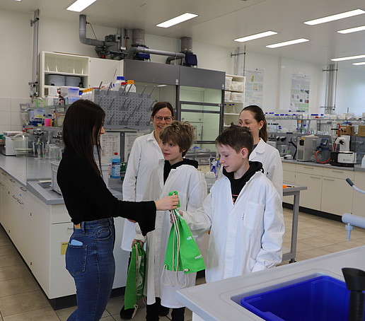 Jonas und Béla bekommen von einer Studentischen Hilfskraft im Labor für pharmazeutische Biotechnologie einen grünen Hochschulsportbeutel überreicht.