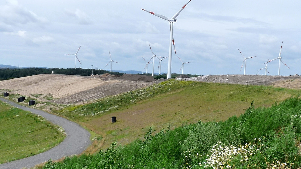 Waste management in practice. Exterior view of the Wetro landfill site. Green hills and cleared areas in the foreground, wind turbines in the background.