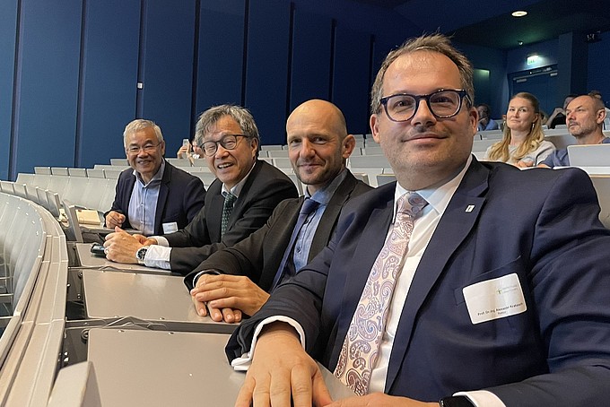 Vier Männer sitzen in einem auditoriumähnlichen Raum an einem Tisch. Prof. Dr. Alexander Kratzsch lächelt in einem dunklen Anzug mit heller Krawatte. Weitere Personen sind im Hintergrund sichtbar.