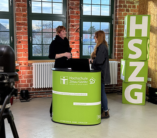 A contributor and the presenter stand behind an HSZG counter in the studio. On the left in the foreground is a black camera. Four HSZG cubes are piled up to the right of the counter.