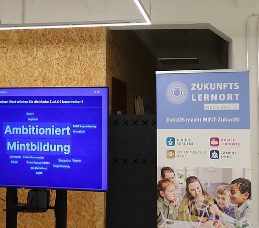 A screen with a presentation stands in front of a cork wall, next to it a ZUKLOS roll-up.