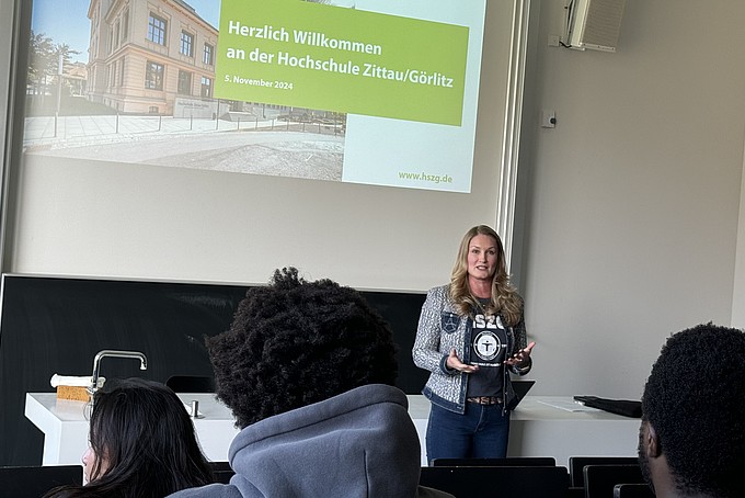 Inna Hauf stands at the front of a lecture hall and speaks to a group of international prospective students from the Rahn Studienkolleg Leipzig. In the background is a screen showing a presentation entitled "Welcome to Zittau/Görlitz University of Applied Sciences". The audience sits in rows in front of her and listens attentively to her greeting.