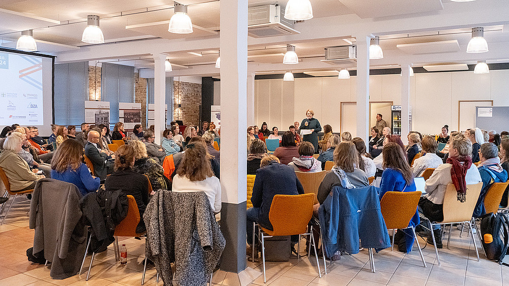 In a large conference room, people sit on orange chairs and listen to a blonde lecturer.