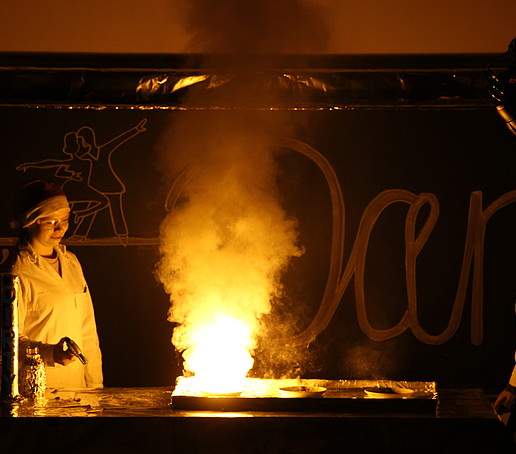 Im Dunkeln führen zwei Personen mit Weihnachtsmütze ein flammendes Experiment auf dem Podium durch.