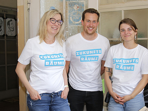 Three people wearing uniform T-shirts with the words "Zukunftsräume" (future spaces) are standing in a fragrance seminar room.