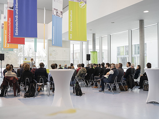 Rows of chairs and high tables are set up in the foyer of House Z IV. The audience seated there listens to a speaker.