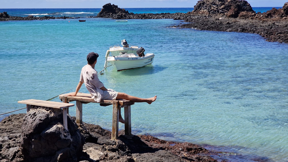 A young man sits on a bench on the beach. A white boat floats in the bay on the azure blue water in front of him.