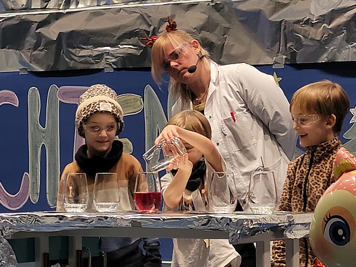 Three children experiment with test tubes and jars of different colored liquids on stage. A laboratory assistant stands in the background.