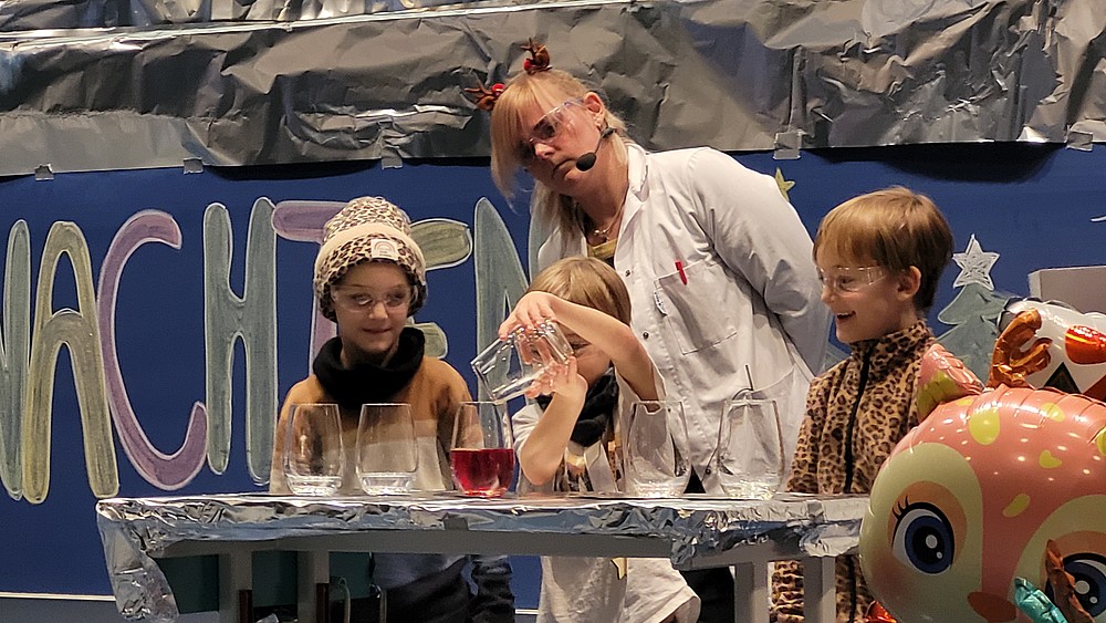 Three children experiment with test tubes and jars of different colored liquids on stage. A laboratory assistant stands in the background.