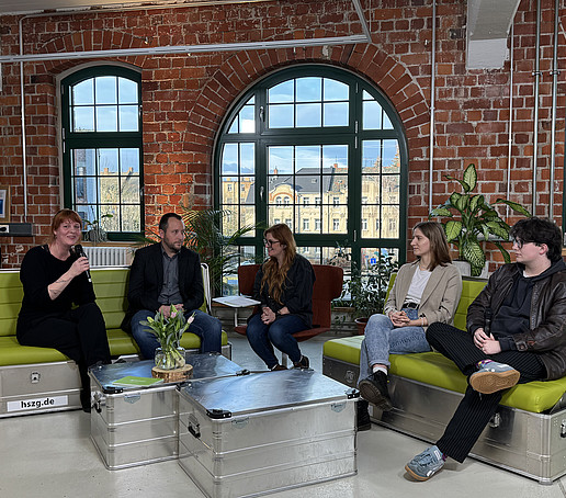 In a studio with brick walls and large windows in the background, participants sit on green sofas and chat.