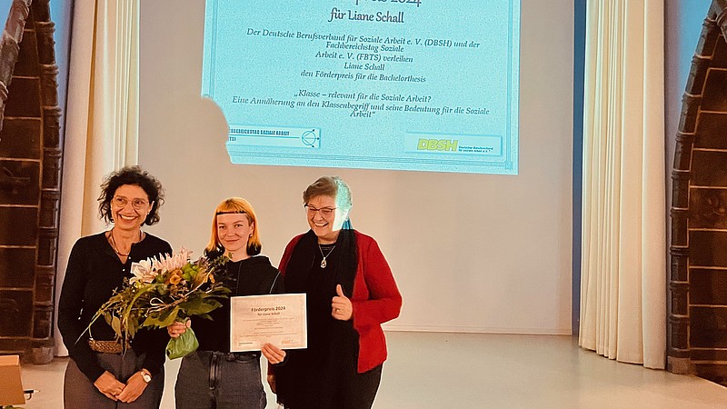 Drei Frauen stehen vor einer Beamerleinwand. In der Mitte steht die Preisträgerin mit ihrer Urkunde.