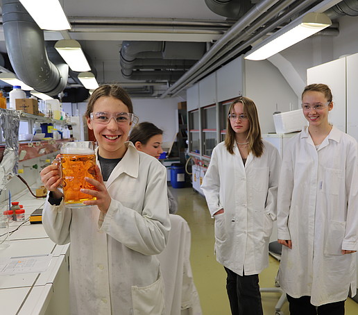 Eine Schülerin hält einen Bierkrug mit einer orangenen Flüssigkeit mit weißem Schaum in der Hand und lächelt in die Kamera.