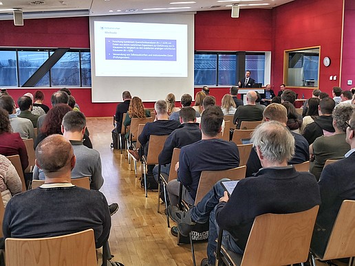 In einem gut gefüllten Seminarsaal mit roten Wänden hält ein Mann einen Vortrag neben einer Leinwand, auf der die Präsentation abgebildet ist.