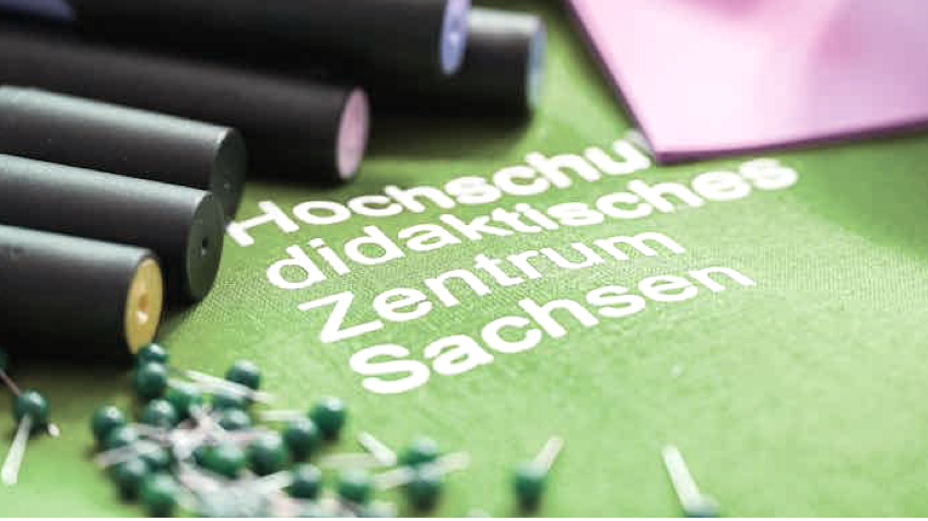 A green printout with the inscription Hochschuldidaktisches Zentrum Sachsen lies on a table surrounded by pins.