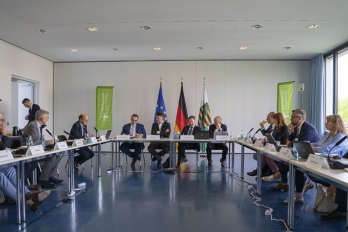 Participants in the cabinet meeting sit in a semi-circle with 'Hochschule' banners in the background.