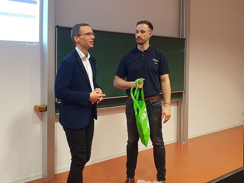 Prof. Kornhuber and Mr. Schumann are standing in front of the blackboard in the lecture hall. Mr. Schumann holds a green bag in his hand while Prof. Kornhuber speaks.