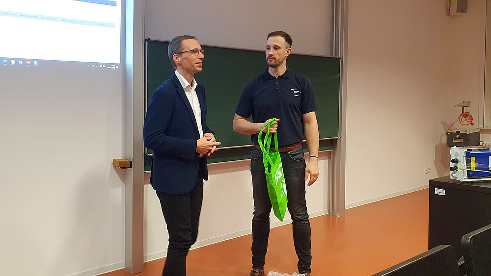 Prof. Kornhuber and Mr. Schumann are standing in front of the blackboard in the lecture hall. Mr. Schumann holds a green bag in his hand while Prof. Kornhuber speaks.