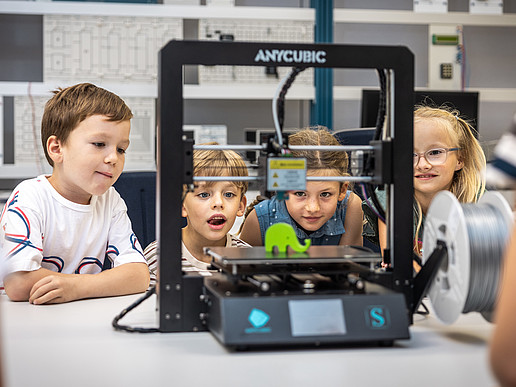 Vier Kinder sitzen im Elektrotechnik-Laborsaal und schauen sich einen kleinen, grünen Elefanten auf einer Plattform eines Gerätes an.