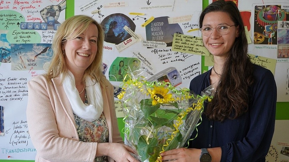 The Vice-Rector and Tanja Schwarz hold a bouquet of flowers between their hands in front of a pinboard.