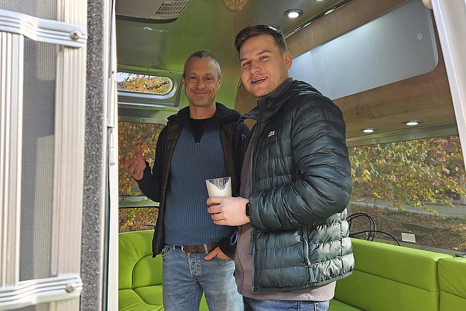 Two guests are standing in front of the green sofa in the trailer.