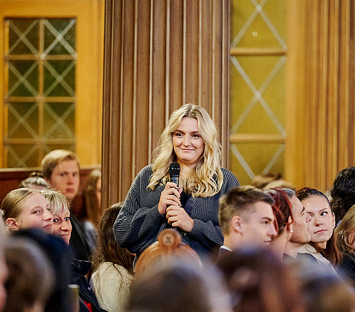 A student is holding a microphone in her hand and has risen from a full row of seats.