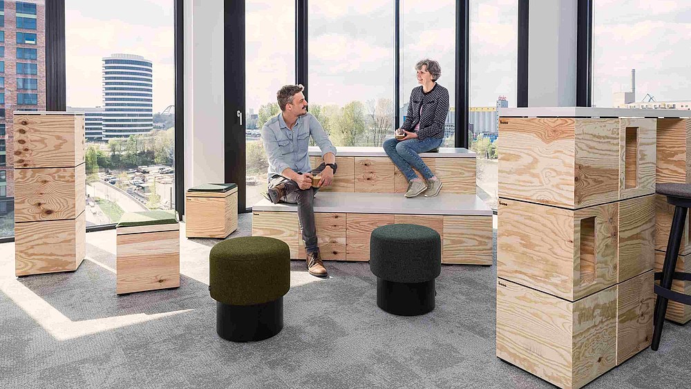 An advertising photo of two people talking while sitting on modern furniture in an office building with large windows in the background.