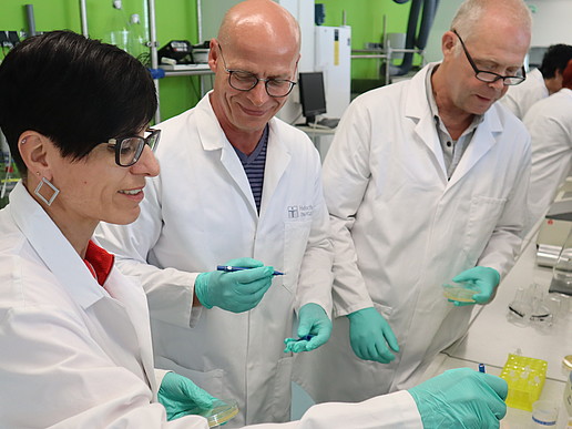 Im Labor hält eine Biologielehrerin eine Pipette in der Hand. Ihre Kollegen schauen dem Experiment zu.